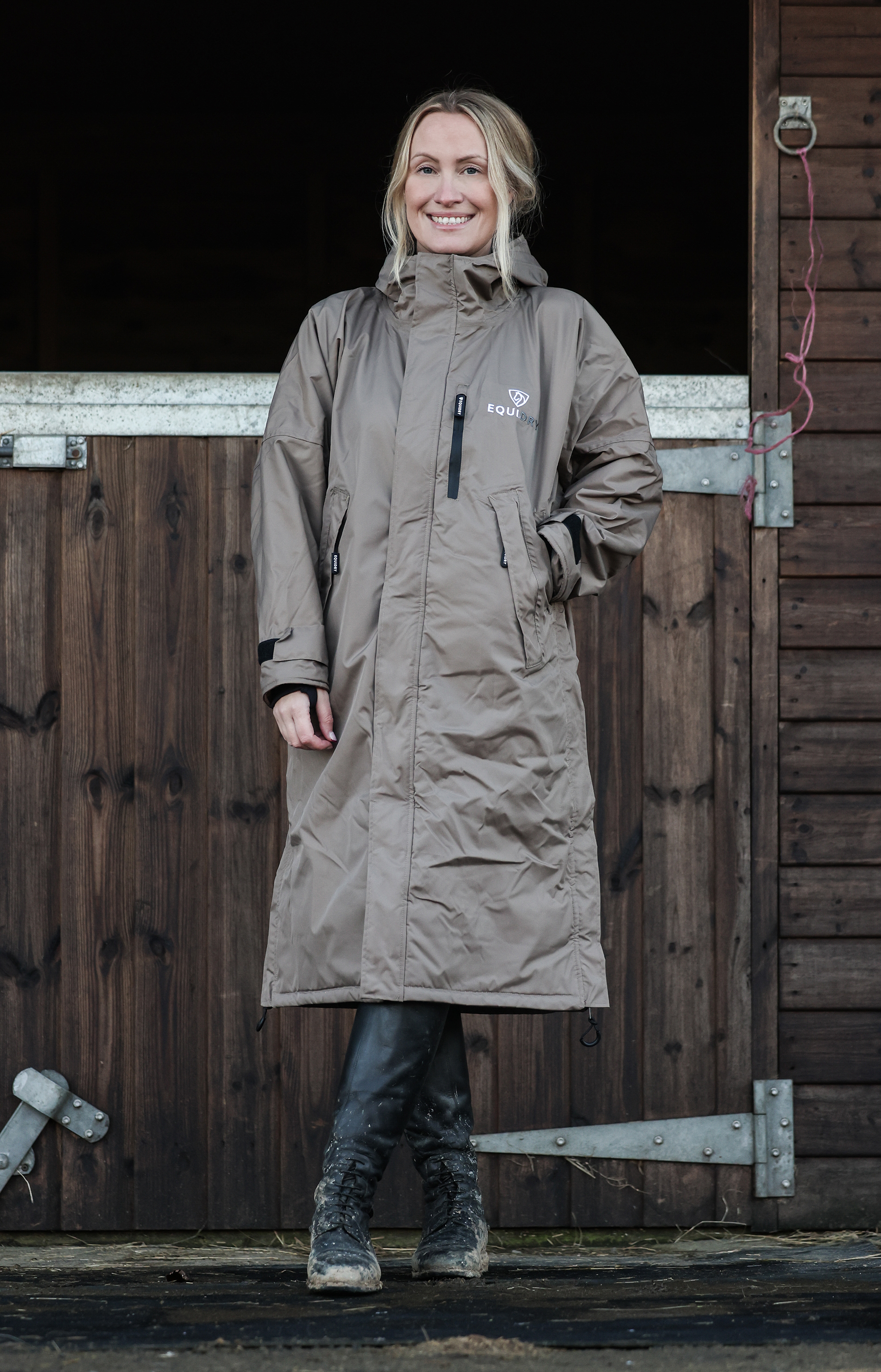 EQUIDRY Evolution Women's Fleece Lined Waterproof Coat - Taupe. Woman in equestrian coat standing by barn door.